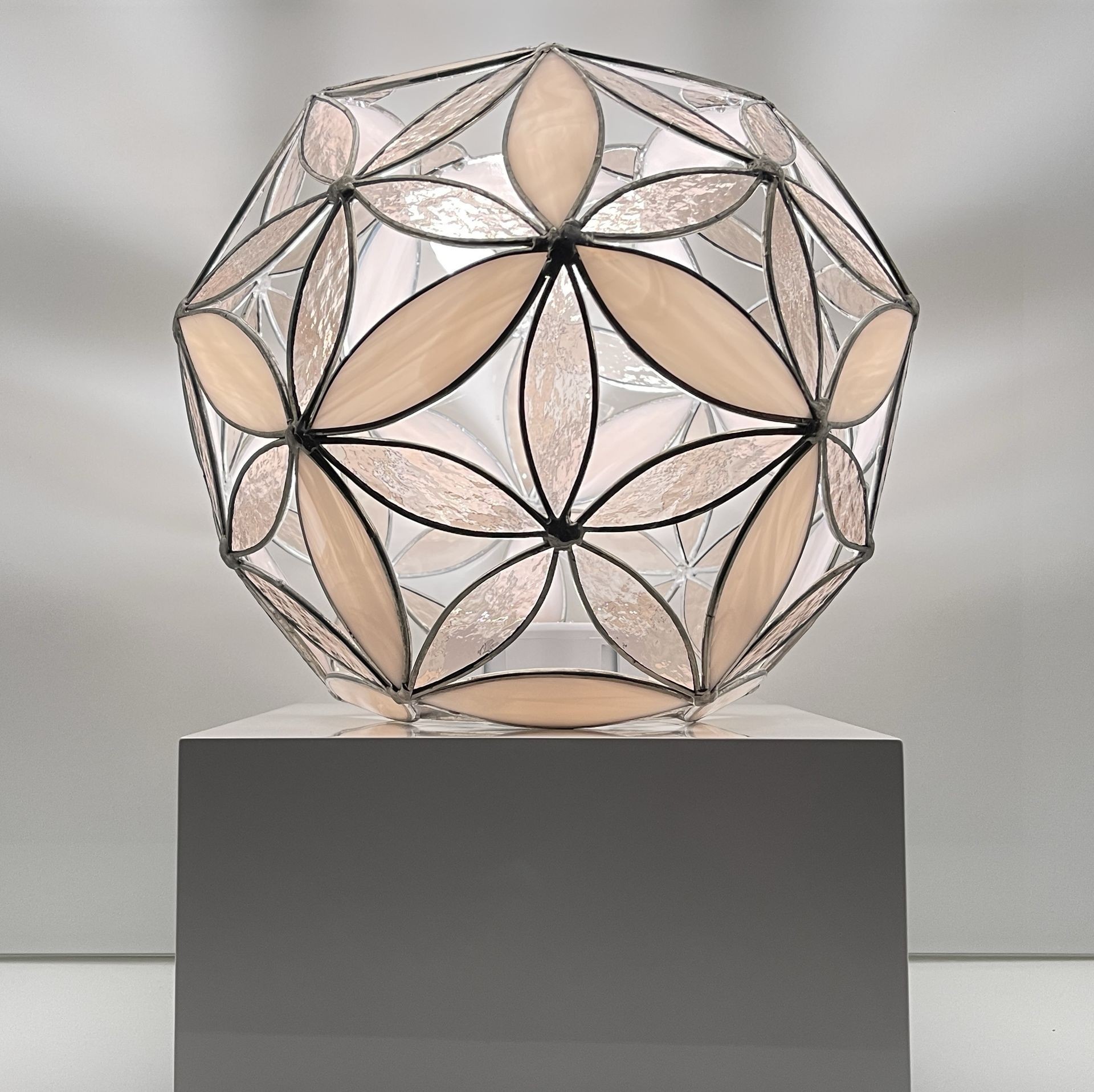 Geometric sculpture with floral stained glass pattern on a pedestal, featuring beige and clear panels.